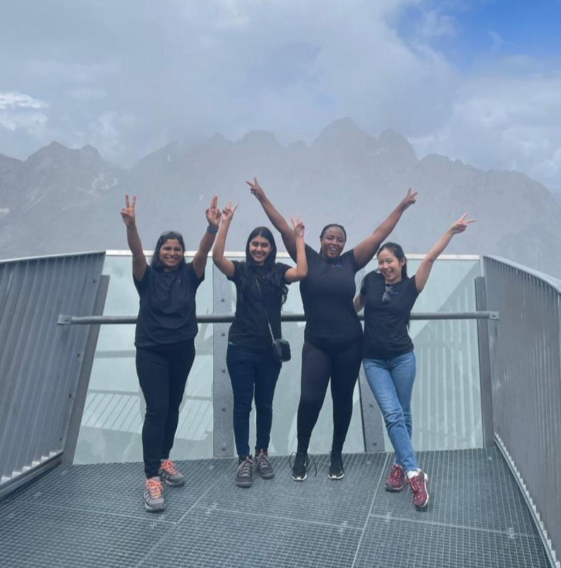 Kavita, Sumedha, Eva and Sara stand on a platform high in the Alps, smiling and raising their arms in celebration against a backdrop of misty mountains and cloudy skies.